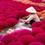 High angle of anonymous woman in traditional incense sticks sitting among rows of bright incense sticks in Vietnam village in daylight