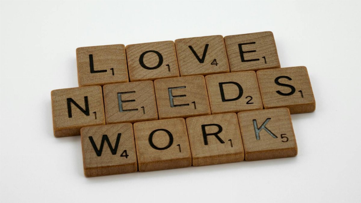 Scrabble tiles spelling 'Love Needs Work' on a white background, symbolizing relationship effort.