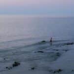 A serene twilight scene with a lone fisherman wading in the tranquil sea waters.