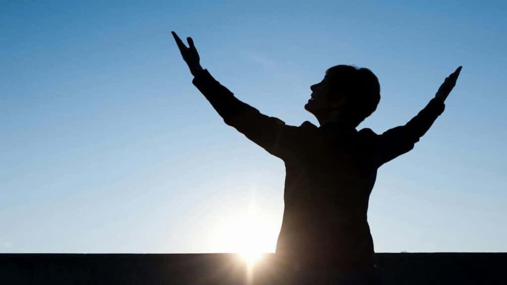 Silhouette of a person raising hands against a sunrise with a clear blue sky, exuding a sense of freedom.