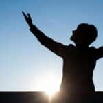 Silhouette of a person raising hands against a sunrise with a clear blue sky, exuding a sense of freedom.
