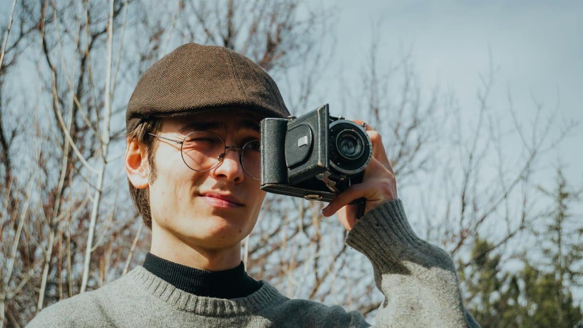 Young man outdoors holding a vintage camera, wearing a flat cap and eyeglasses.