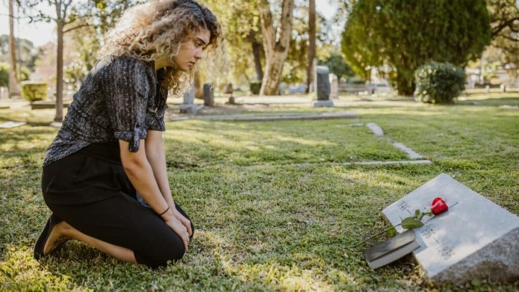A woman kneeling by a grave in a sunlit cemetery, expressing grief and remembrance.