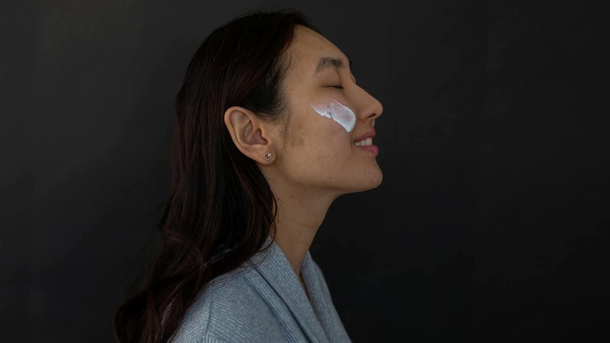 Asian woman in profile with eyes closed, enjoying a skincare routine with cream on her cheek.