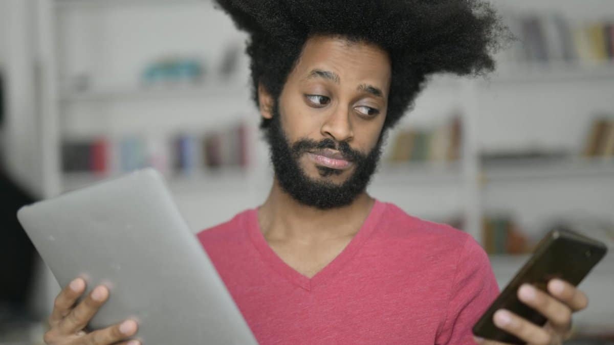 A man with afro hair multitasking with a tablet and smartphone indoors, showcasing technology use.