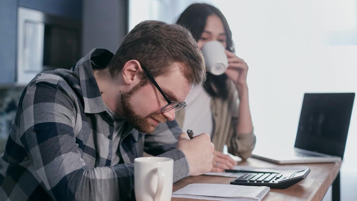 A couple sits at a kitchen table, reviewing finances with a laptop and calculator.