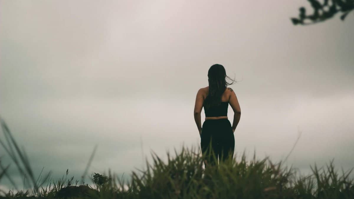 Silhouetted woman standing in a grassy field under a cloudy twilight sky, evoking solitude