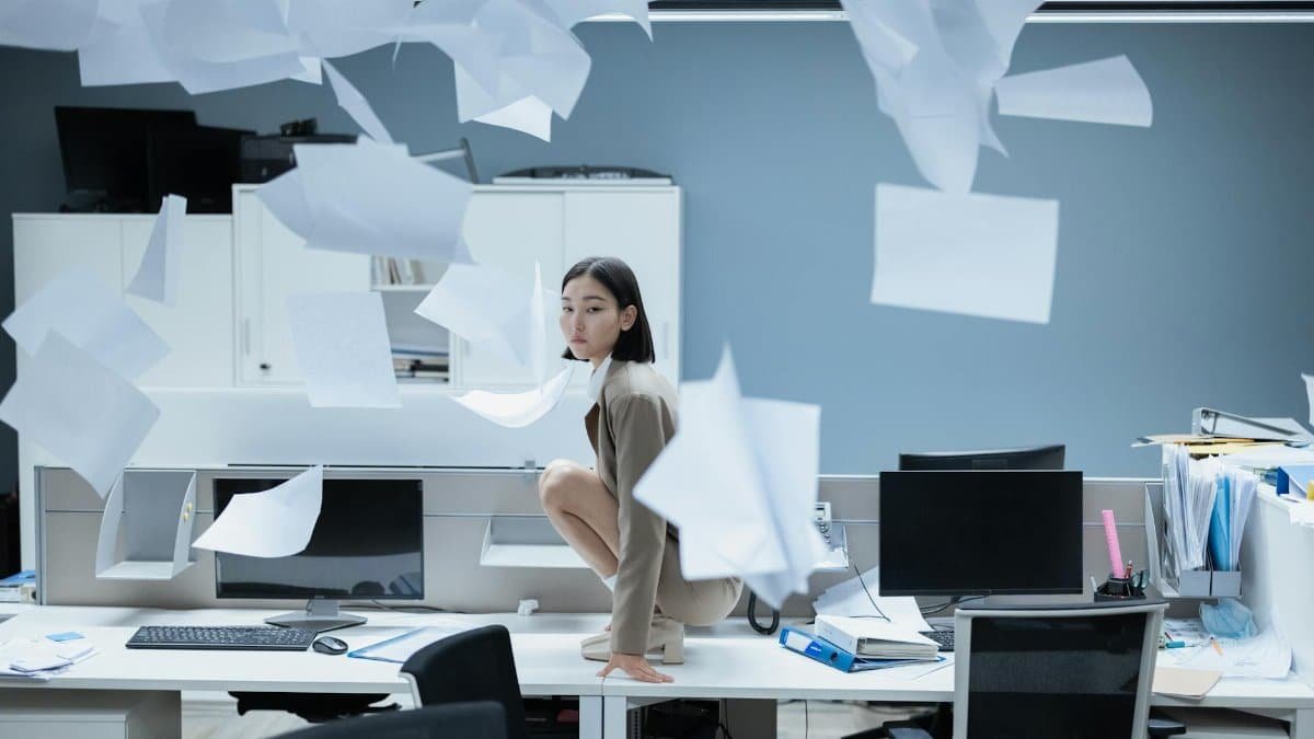 Woman perched on a desk in a chaotic office with papers flying, representing workplace stress.