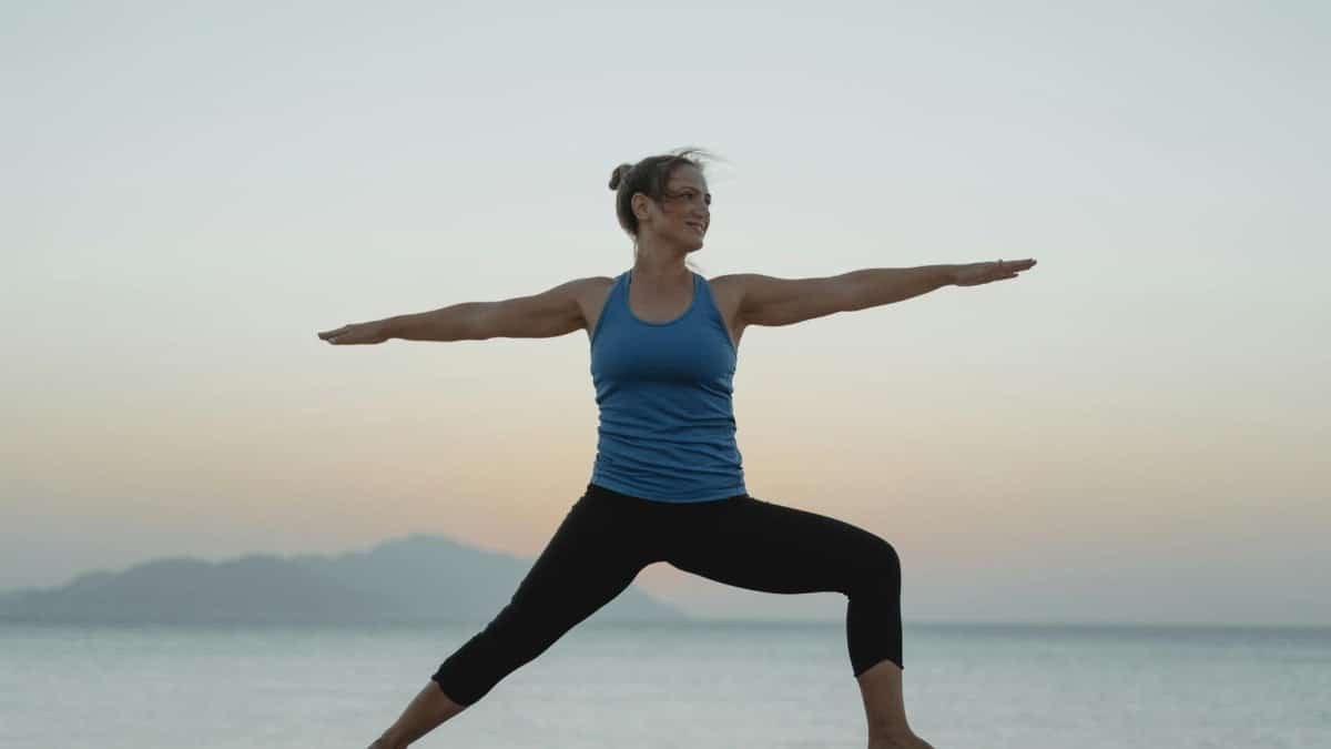 Adult woman practicing yoga on a sandy beach during sunrise with calm sea in the background.
