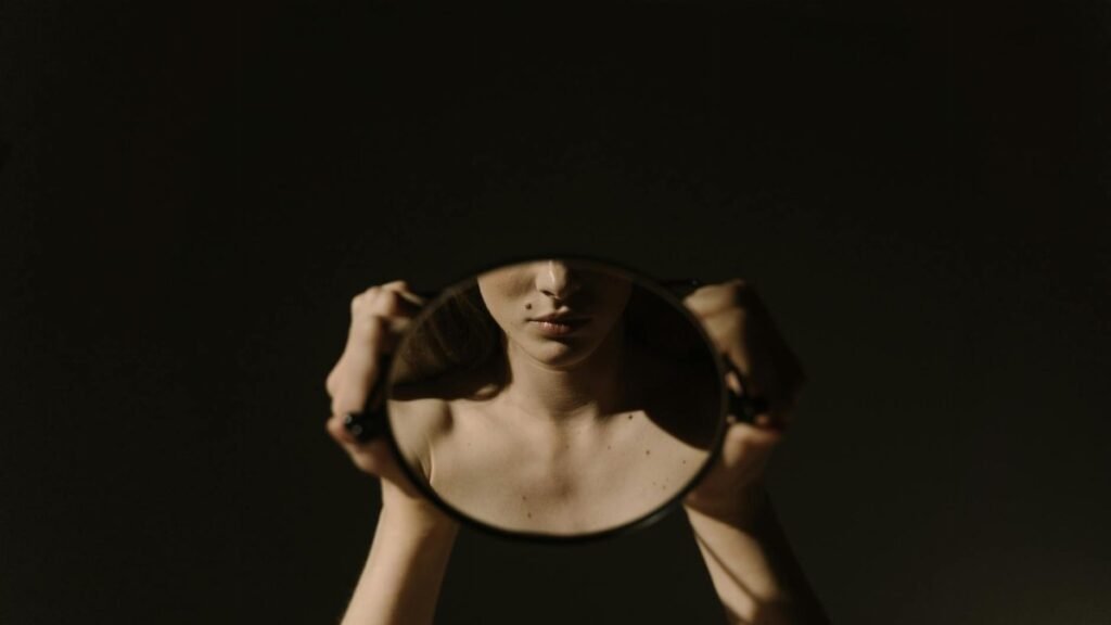 Artistic portrait of a woman holding a mirror, reflecting her face, with a dark, moody background.