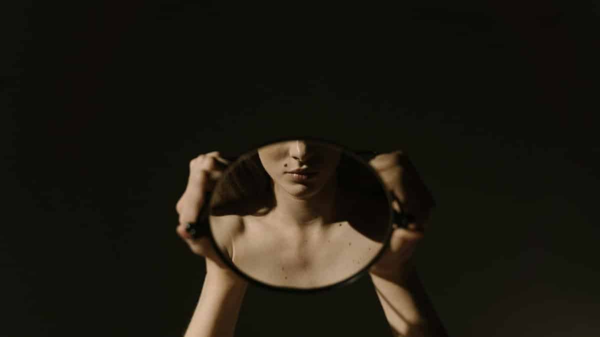 Artistic portrait of a woman holding a mirror, reflecting her face, with a dark, moody background.