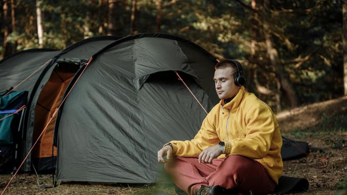 A man enjoys quiet meditation outside a tent in a forest, wearing headphones and a yellow jacket.