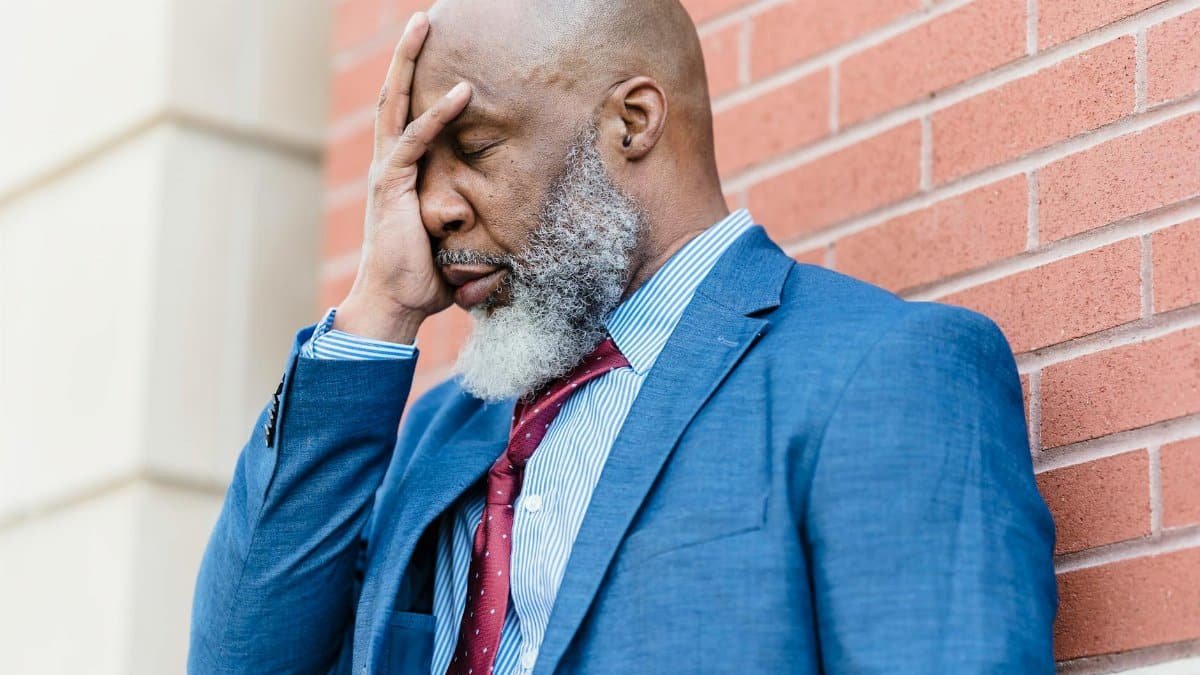 A bearded man in a suit showing emotions while leaning against a wall.