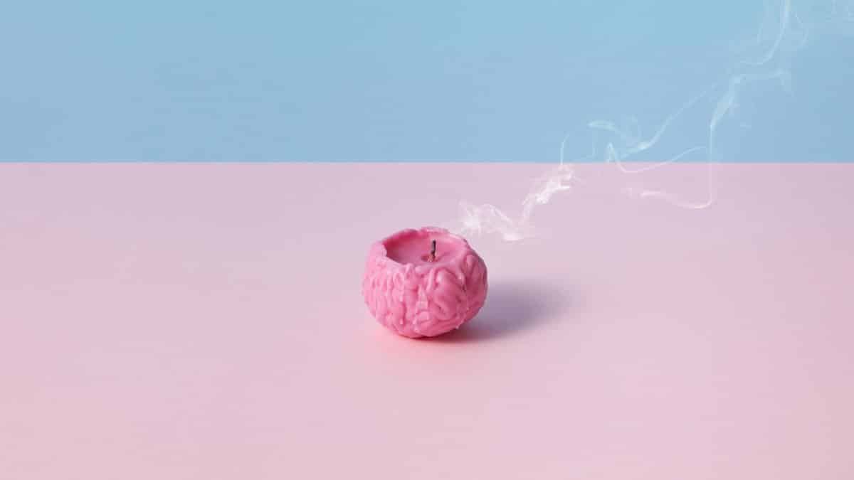 Conceptual image of a pink brain-shaped candle with smoke, on a pastel pink and blue background, symbolizing burnout.