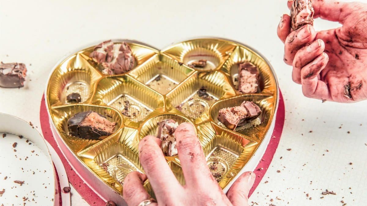 Close-up of messy hands indulging in chocolates from an empty heart-shaped box.