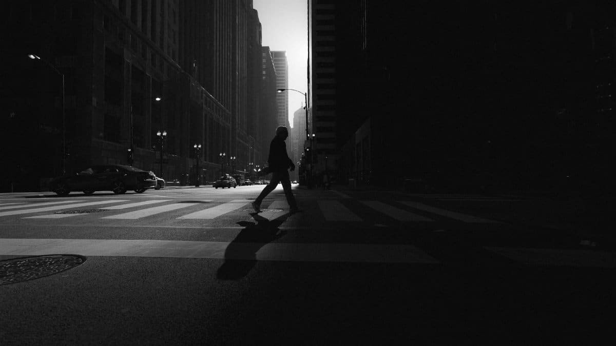 Black and white silhouette of a person crossing a city street with tall buildings lining the roadway.