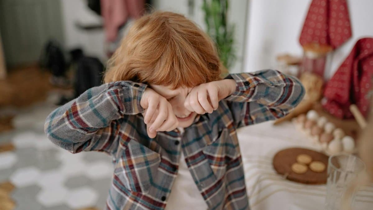 A young boy with red hair wipes his eyes in a cozy kitchen, evoking emotion and childhood innocence.