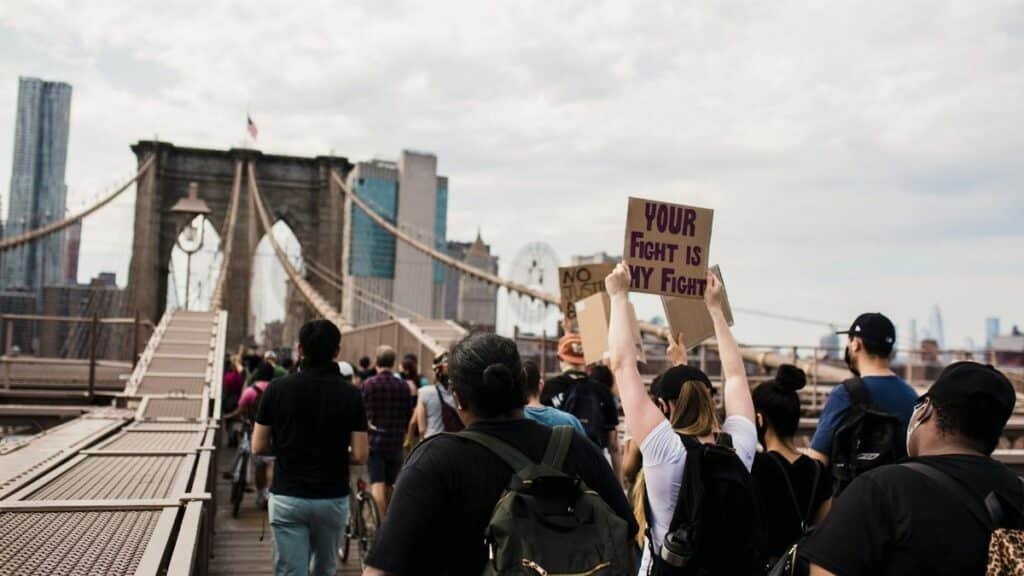 A diverse group of protesters cross Brooklyn Bridge with signs advocating for justice and solidarity.