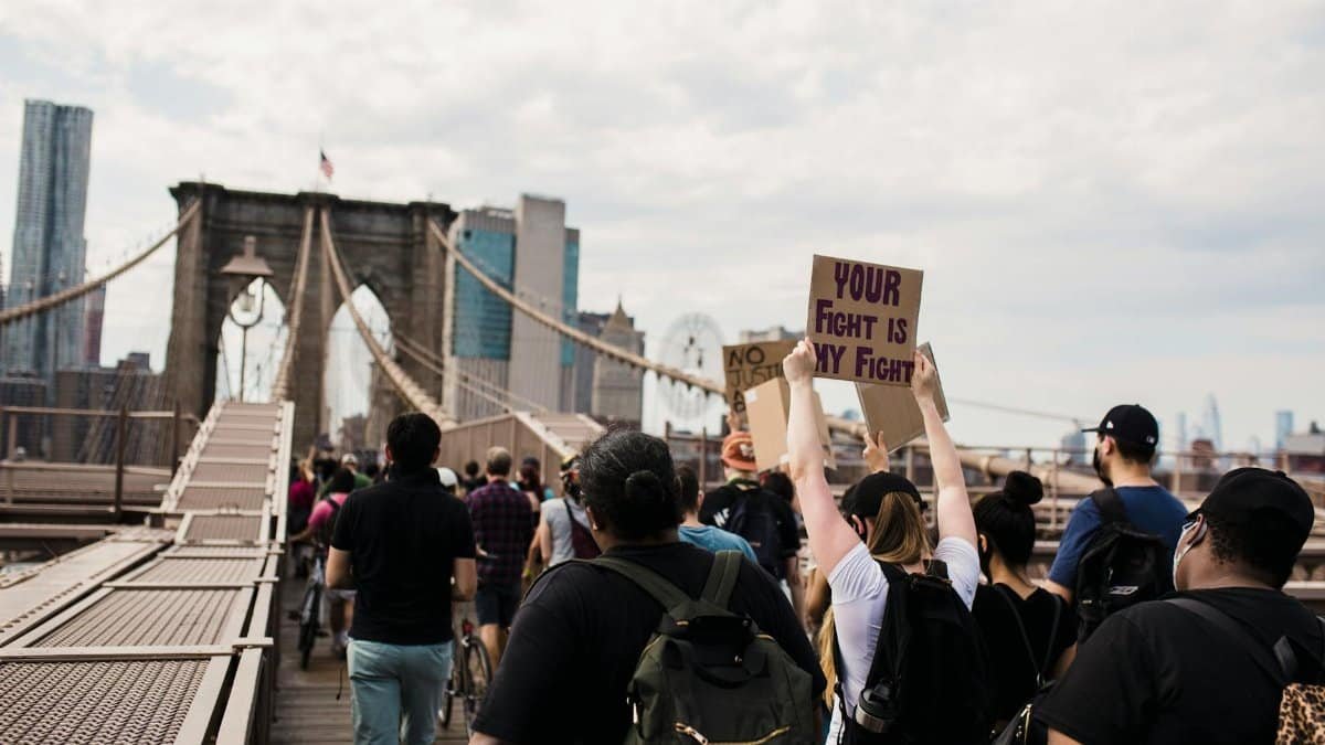 A diverse group of protesters cross Brooklyn Bridge with signs advocating for justice and solidarity.