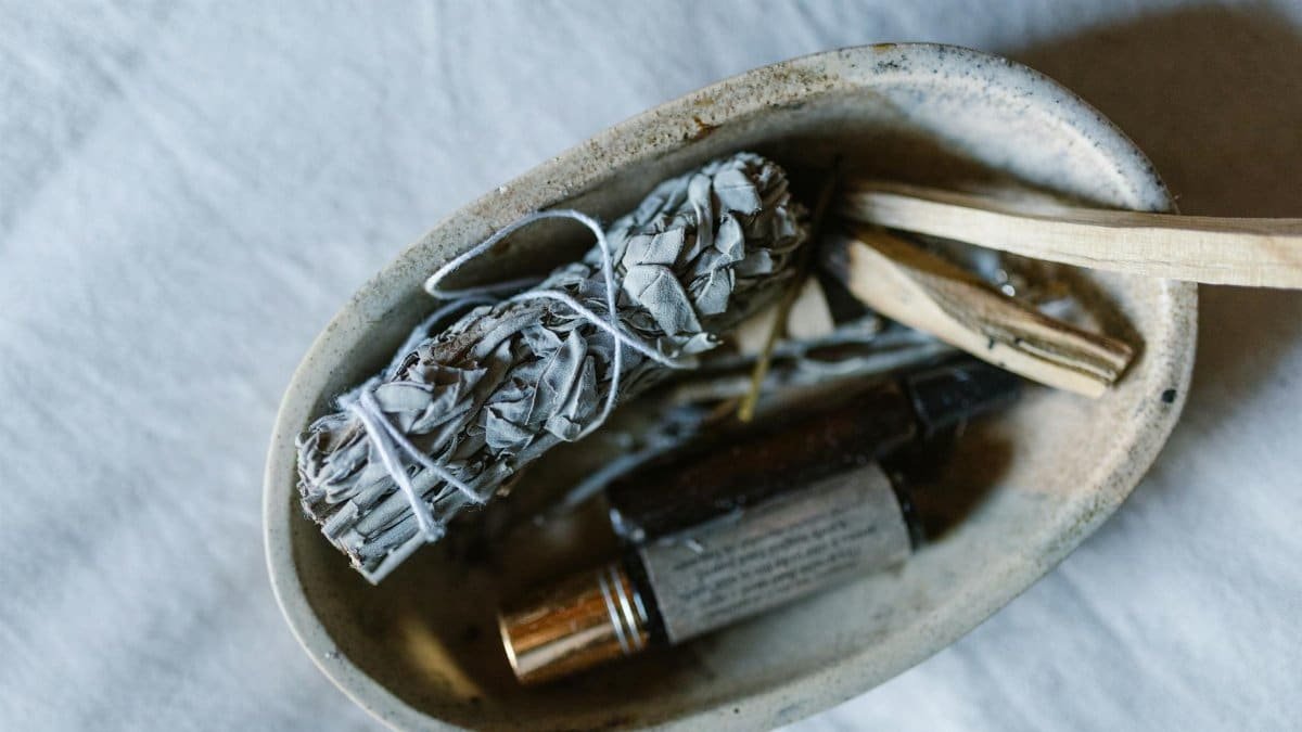 A serene still life featuring a bundle of sage, essential oils, and wooden sticks in a rustic bowl.