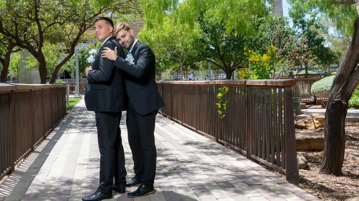 A loving couple in formal suits embracing on a sunny day in a serene park setting.