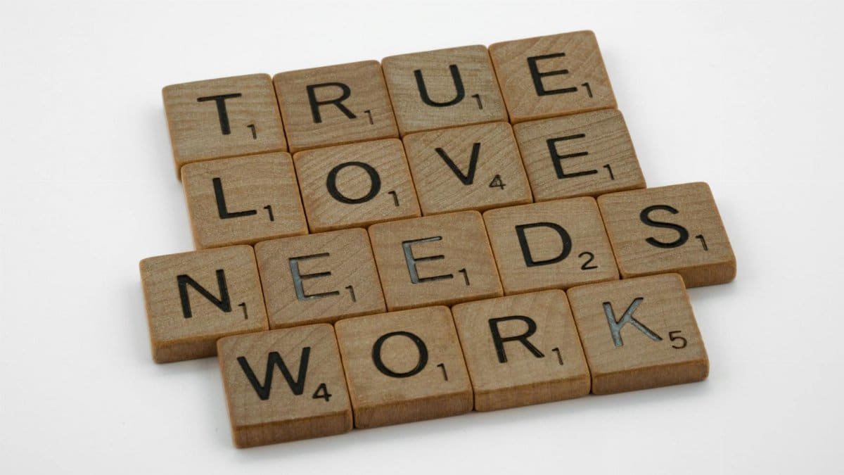 A close-up of wooden Scrabble tiles on a white surface spelling 'True Love Needs Work'.