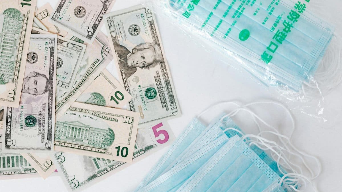 From above composition of stack of USA dollar bills placed near medical protective masks produced in China illustrating concept of medical expenses and deficit during COVID 19