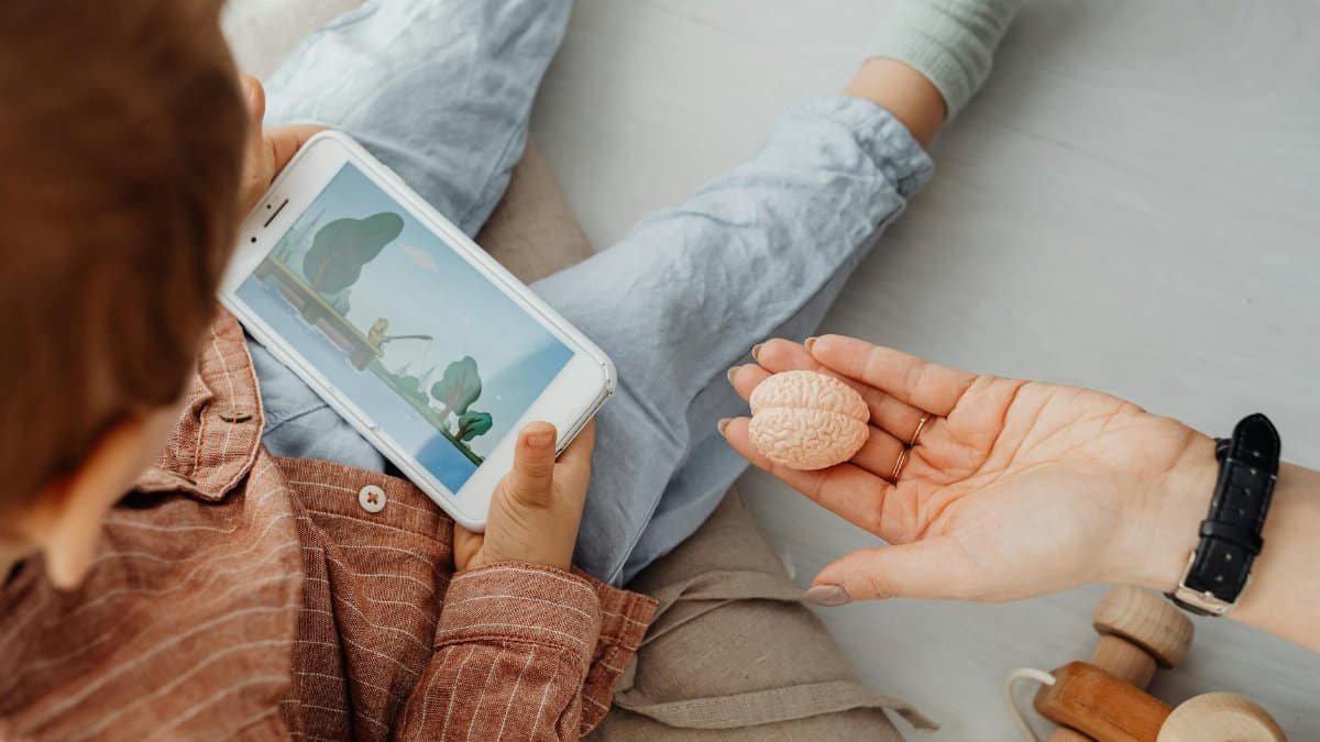 Young child engrossed in a smartphone video while holding a brain model indoors.