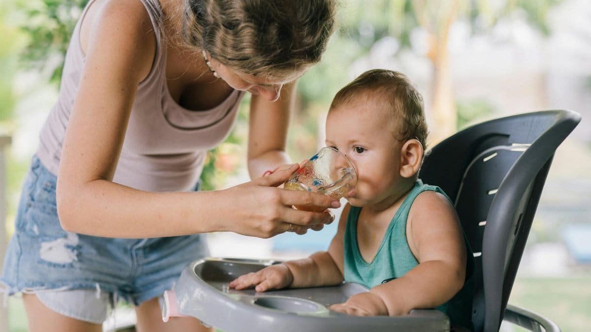 A mother helps her baby drink water from a glass while sitting in a high chair.