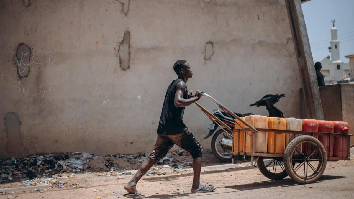 A young man pushes a cart filled with containers down an urban street, showcasing daily life in the city.