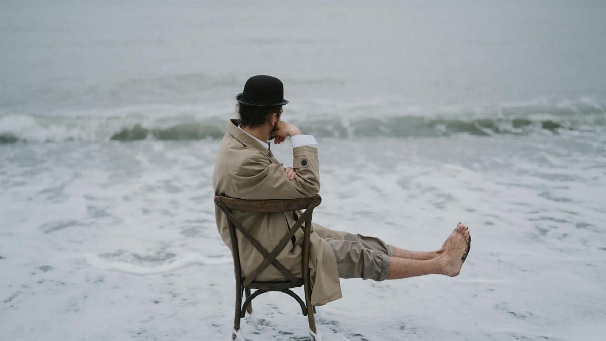 Man in coat and hat sitting on a chair at the edge of the ocean, lost in thought.