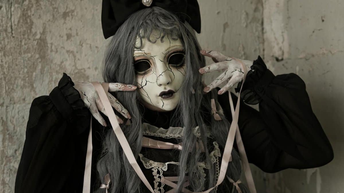 A person in eerie doll-like cosplay with a cracked mask, ribbons, and dark clothing, indoors with dark mood lighting.