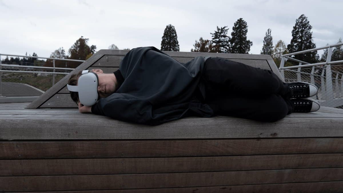 Man lying on a wooden platform outdoors wearing a VR headset. Trees in the background.