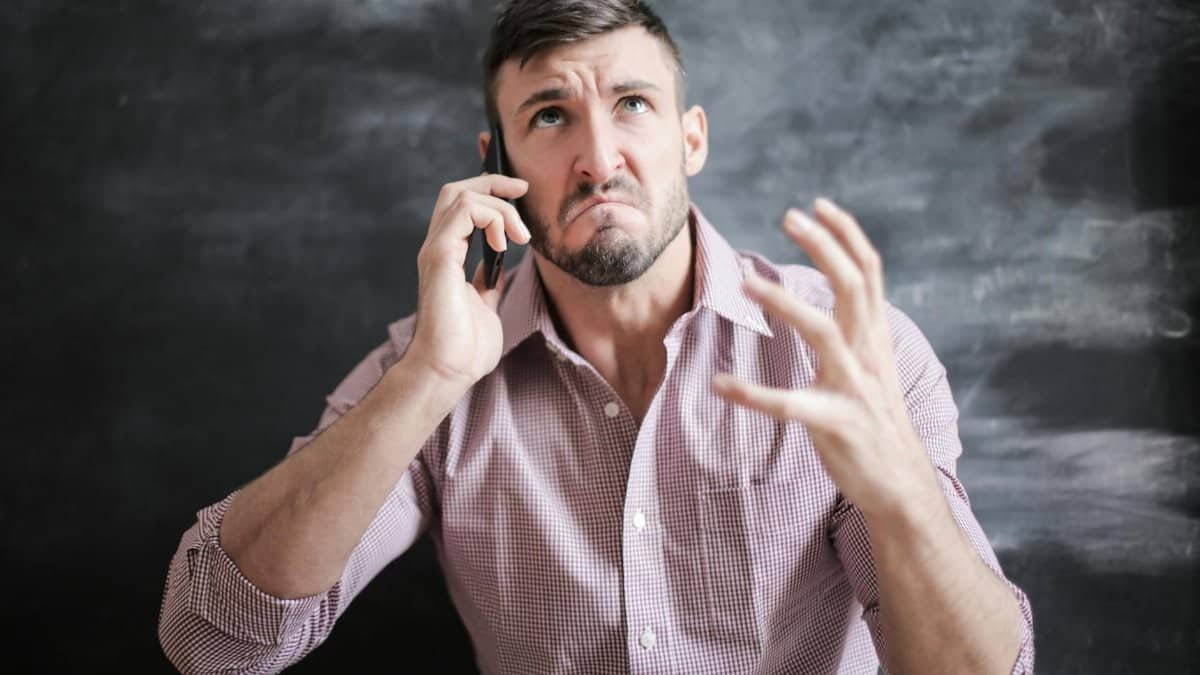 Adult man in pink dress shirt looking stressed while talking on a smartphone indoors.