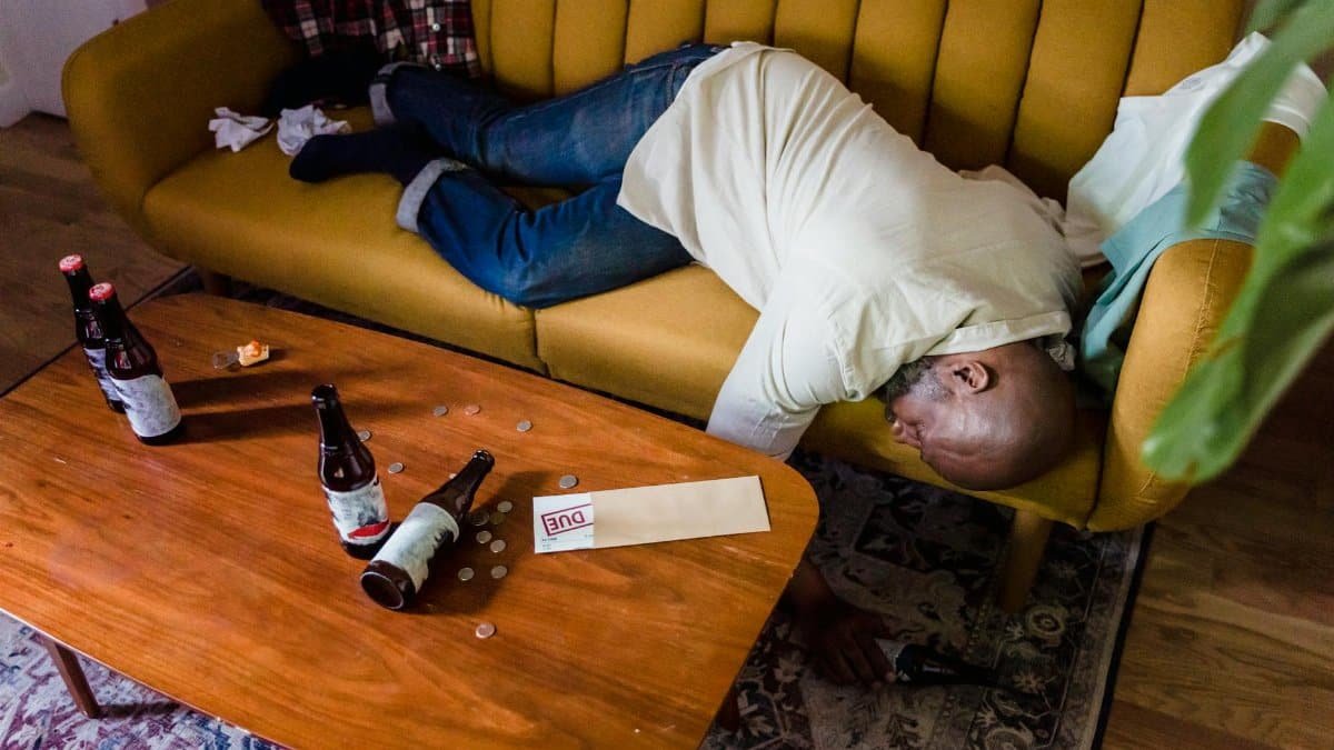 A man sleeping on a couch with empty bottles and an overdue bill, symbolizing financial stress and exhaustion.