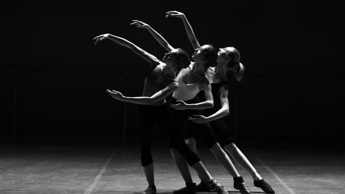 Three female ballerinas perform a synchronized dance on stage in dramatic lighting.