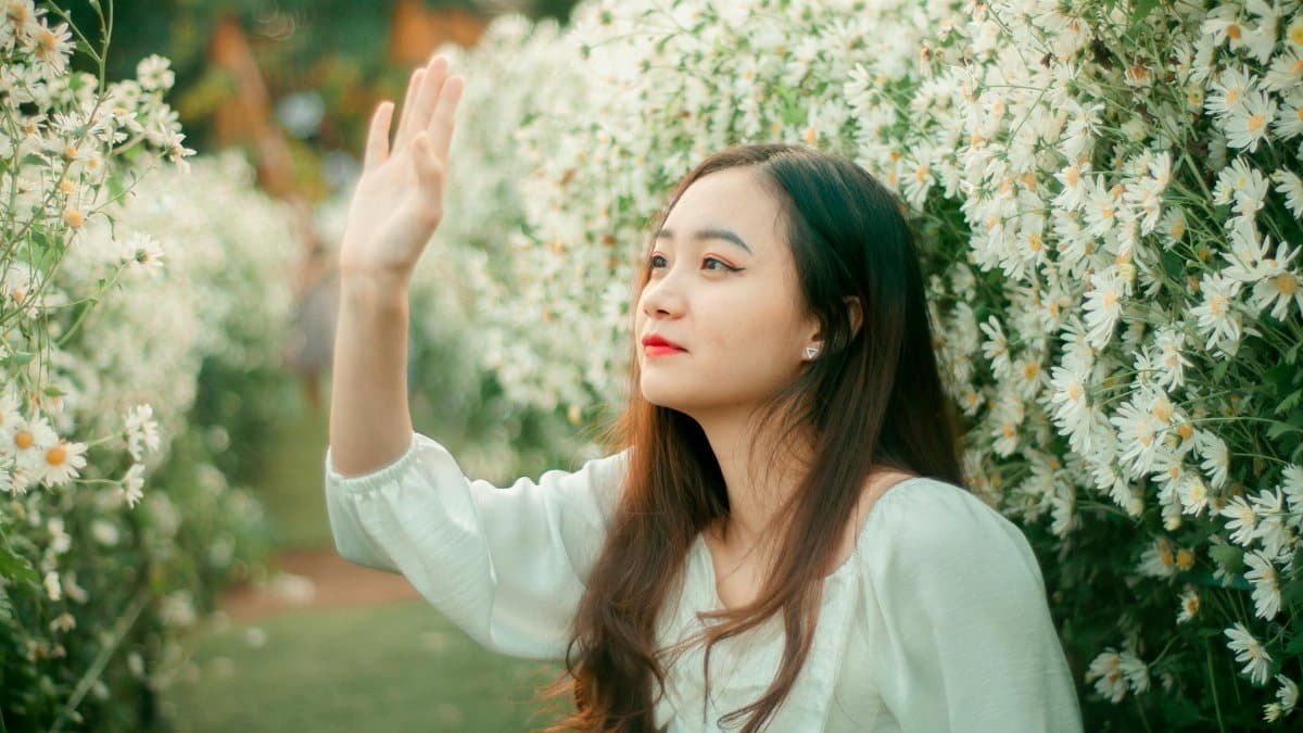 Asian woman with long hair smiling among blooming daisies, waving joyfully.