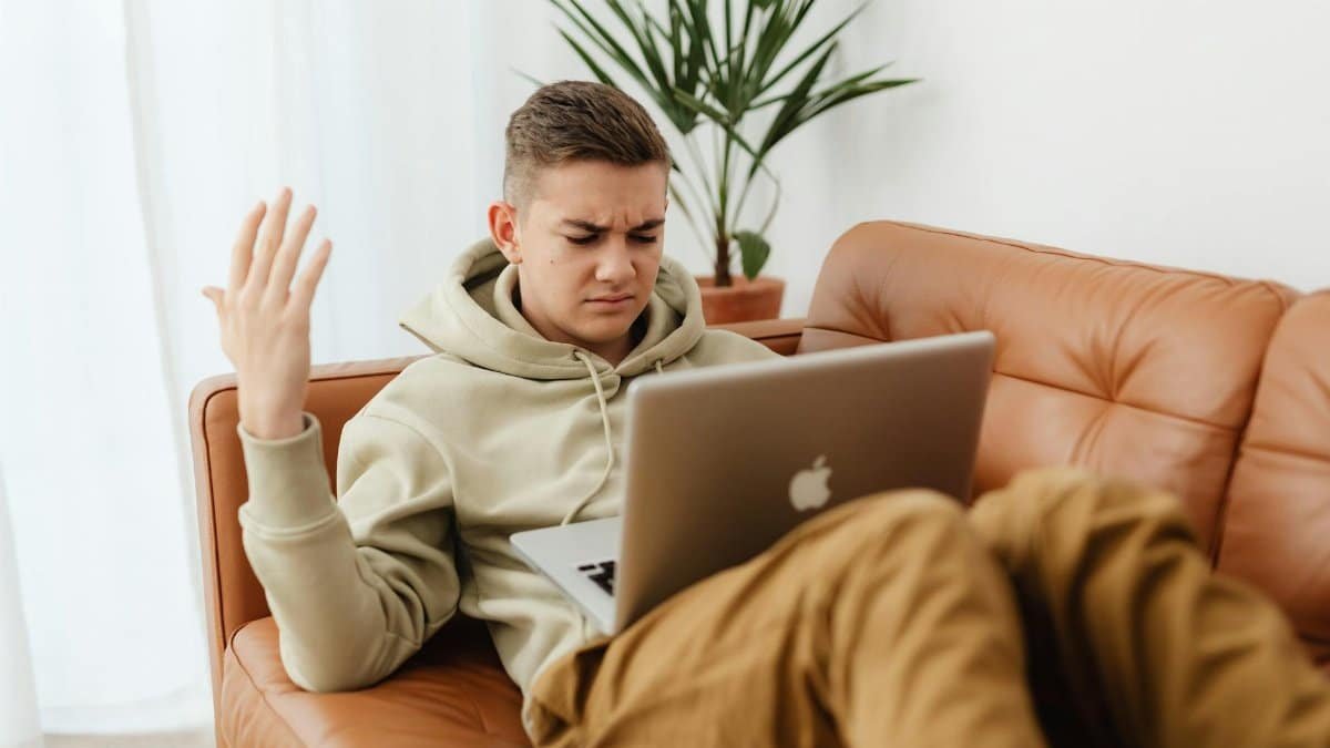 A young man in a hoodie looks irritated while working on a laptop in a cozy indoor setting.
