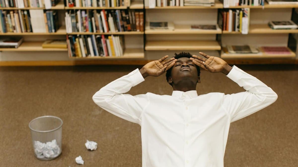 A frustrated man leans back in a library with crumpled papers on the floor.