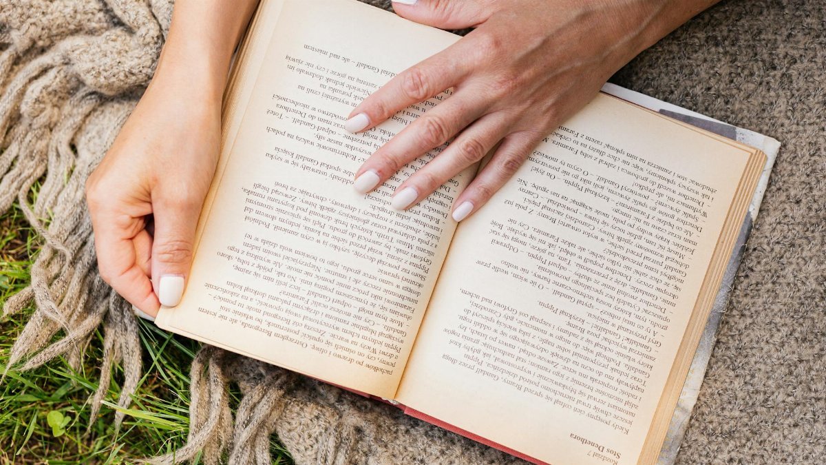Hands with white nails hold an open book outdoors on a blanket, offering a cozy reading moment.