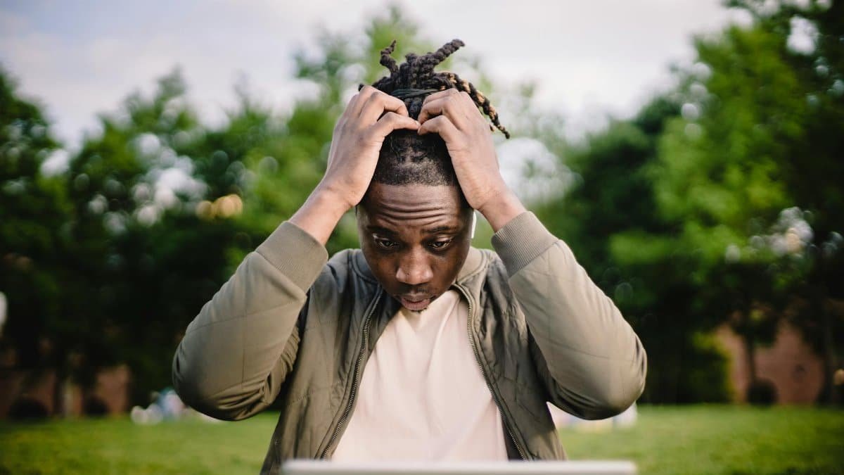 Upset young African American male freelancer clutching head with hands after failure in project while working remotely in park