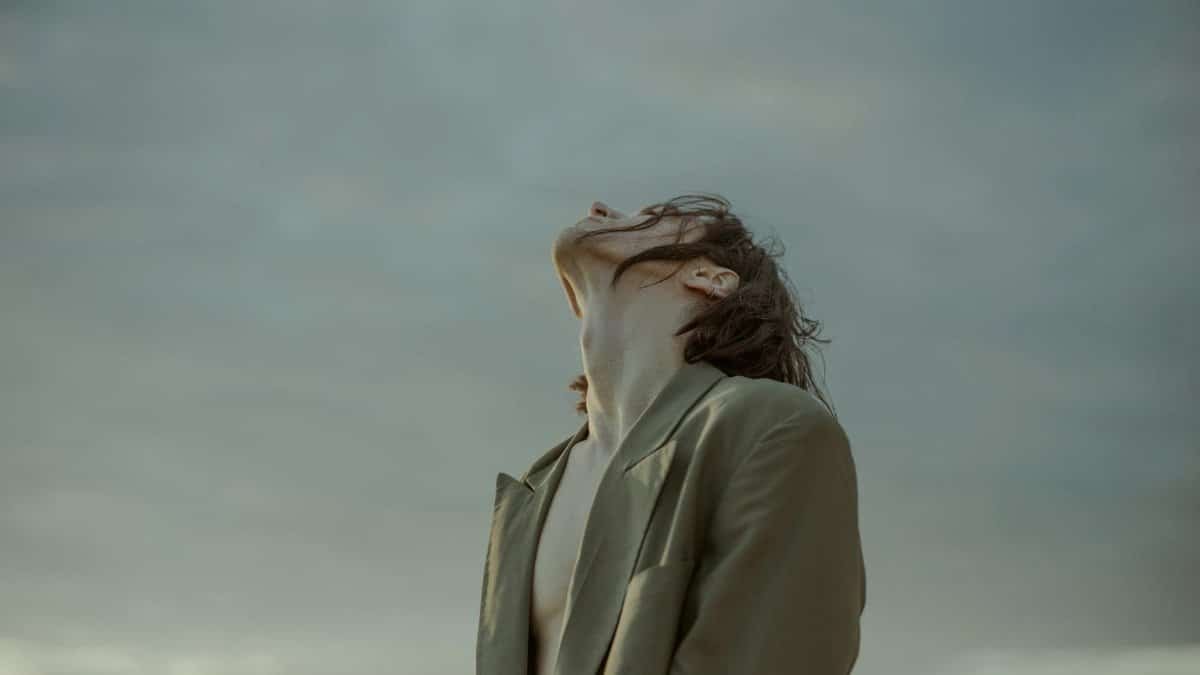 A man in a suit gazes upwards with a cloudy sky background, highlighting fashion and serenity.