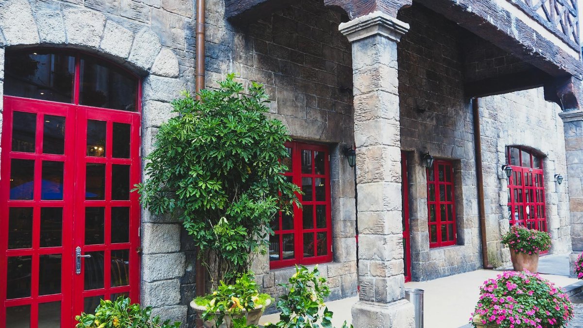 Various fresh floral plants in pots decorating yard of aged stone building with red doors and window in daytime