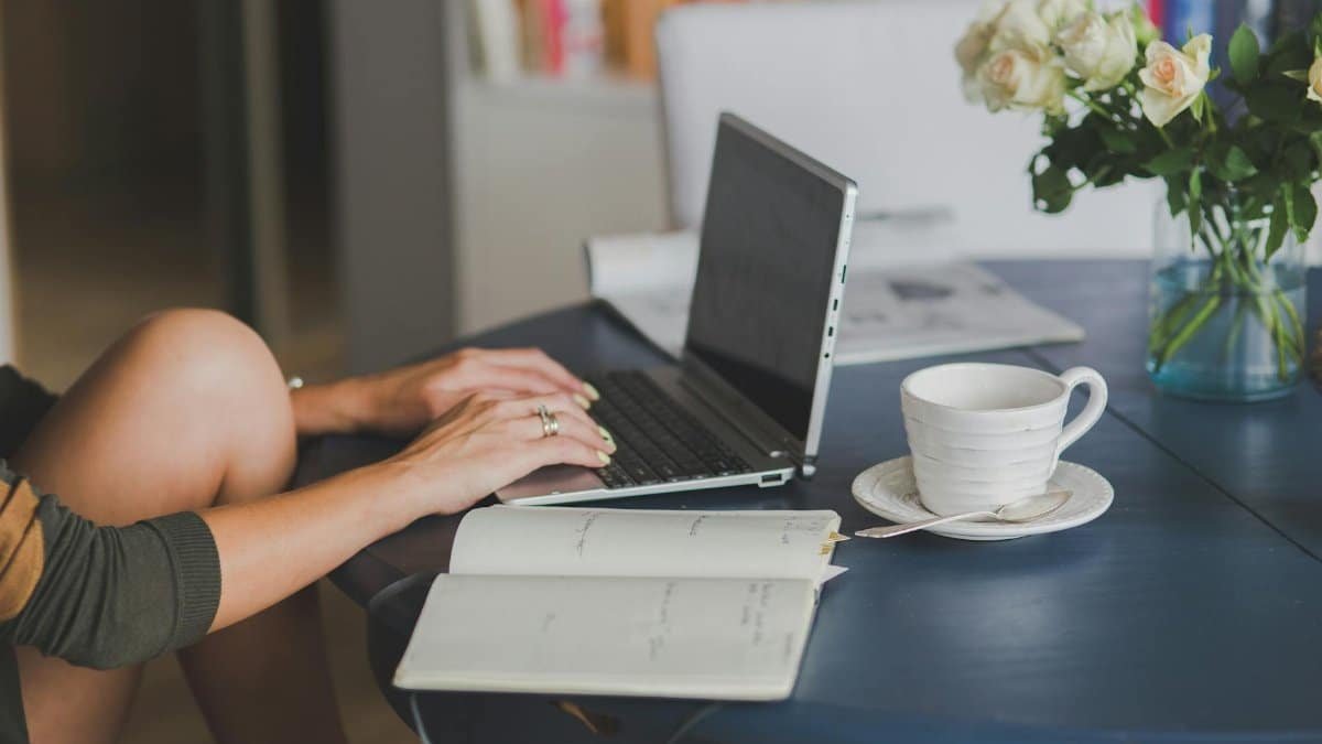 Female freelancer using laptop with coffee at home office desk, surrounded by roses and a planner.