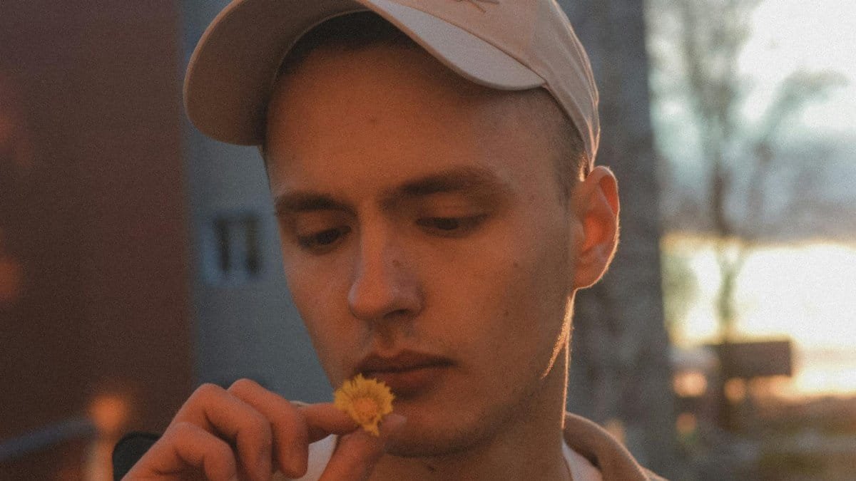 A man in a cap thoughtfully looks at a small yellow flower, capturing a moment of contemplation outdoors.