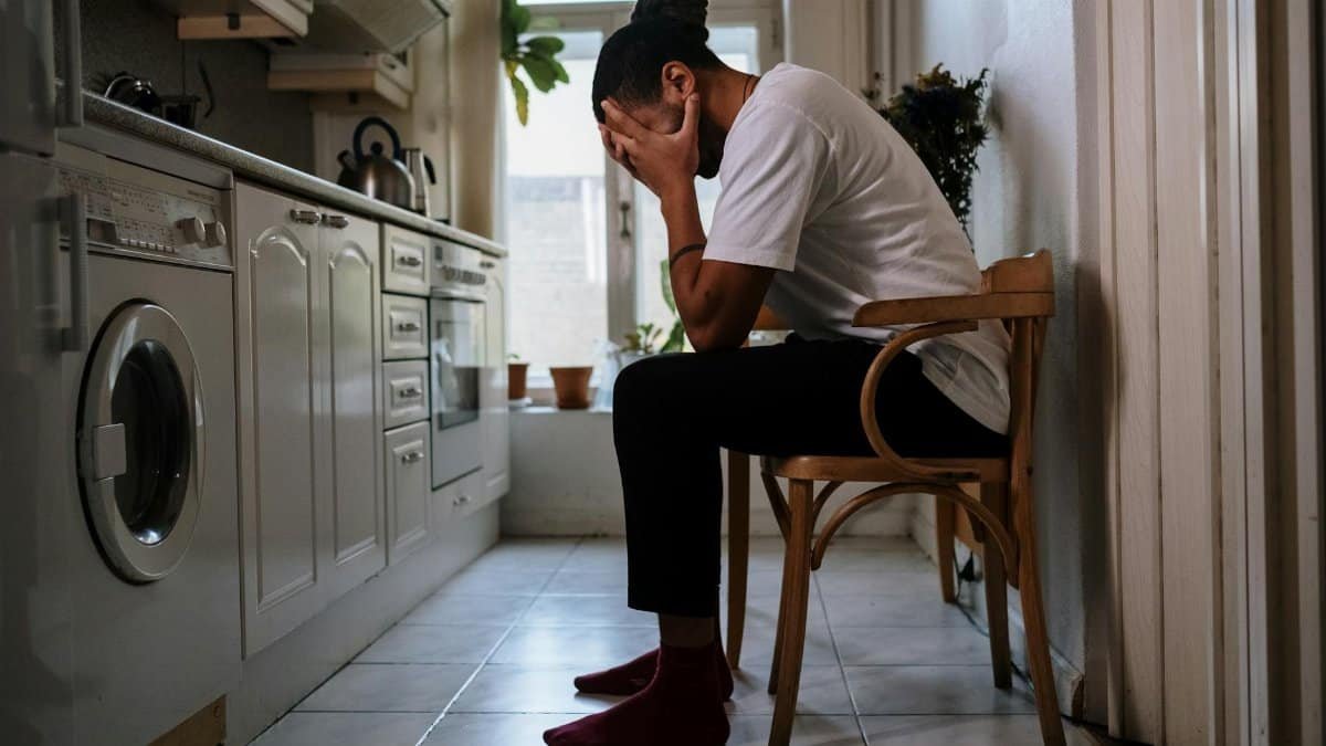 A man sits in a kitchen, visibly stressed with head in hands, capturing an emotional moment indoors.