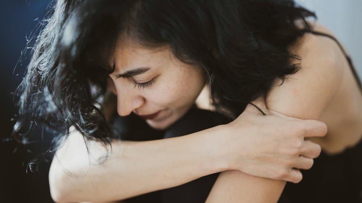 A woman with dark hair appears pensive and emotional, sitting indoors, arms crossed.