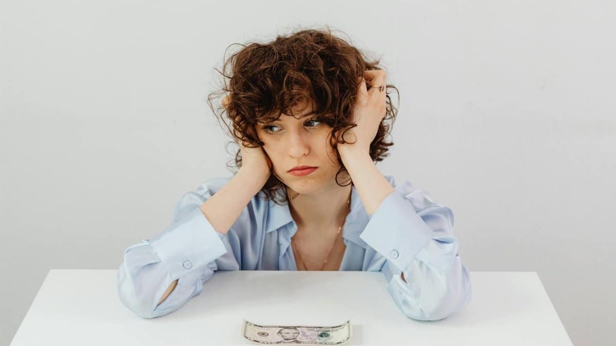A woman in a blue blouse looks worried with a dollar bill on a table, symbolizing financial stress.