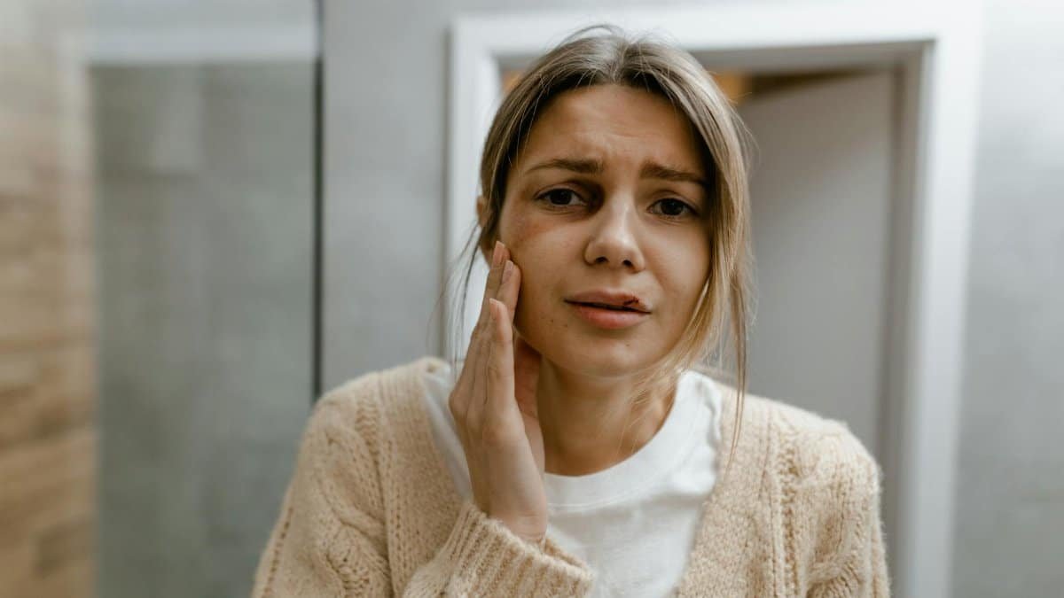 A woman indoors with a worried expression, touching her cheek, in a light-toned room.