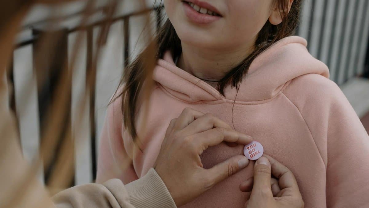 A young girl in a pink hoodie with a motivational 'You Go Girl' badge being pinned on her.